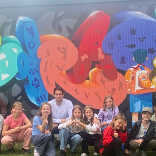 Gruppenfoto der Kinder aus dem Hort der Grundschule Nord gemeinsam mit der KJIV, dem Graffitikünstler und 2 beteiligten Erziehr:innen vor dem Container, auf dem in bunt und mit Bildern der Name Conrad gesprüht wurde.