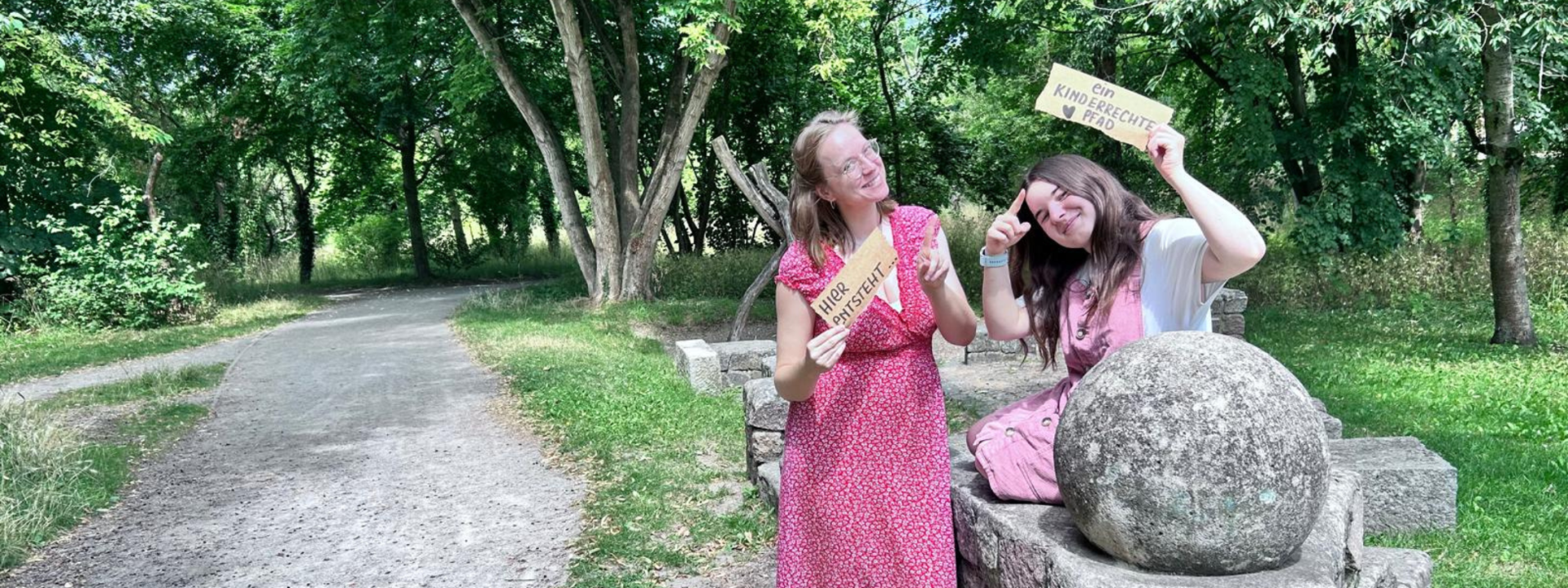 Links ist ein Weg zu sehen (Hartungswall in Stendal). Rechts zwei Personen (Kira und Janine von der Kinder- und Jugendinteressensvertretung), die zwei Schilder hochhalten. Auf den Schildern steht "Hier entsteht..." "ein Kinderrechtepfad